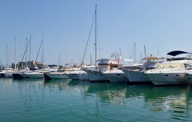 Fototapeta premium A picturesque view of Antibes' Port Vauban, featuring a line of sleek luxury yachts and sailboats docked in the clear blue water. In the distance, the historic Fort Carré stands on the hillside