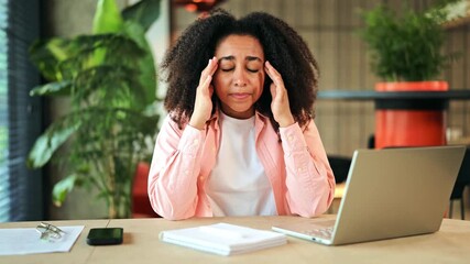 A tired African American businesswoman in a pink shirt experiences a headache while diligently working on her laptop at a desk in a modern office. She feels overwhelmed and stressed. - Powered by Adobe