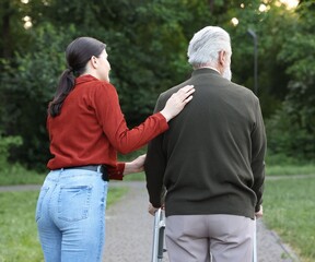 Caregiver supporting elderly man who using walking frame outdoors, back view