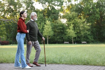 Smiling caregiver supporting elderly man who using walking cane outdoors. Space for text