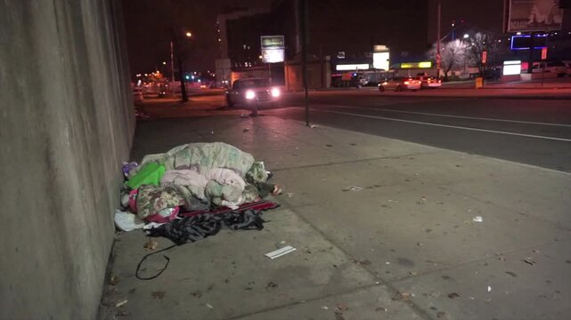 Homeless man sleeps under a pile of blankets on a city sidewalk with blowing trash at night.