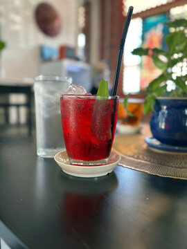A refreshing red drink with ice and a pandan leaf garnish sits on a table, next to a glass of water, in a cozy Malaysian cafe setting.