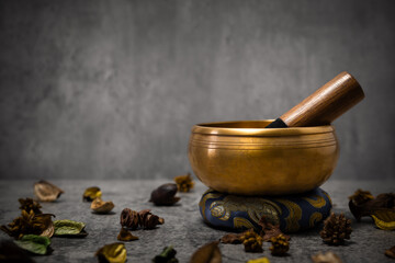Golden tibetan singing bowl standing on a small blue cushion with wooden stick and dried plants scattered around, promoting relaxation and meditation against a gray backdrop