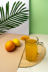 Cold orange juice in a beer stein on a woven coaster. Fresh lemon and orange with a tropical leaf on a modern, colorful background. Healthy lifestyle.