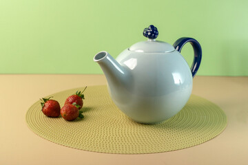 Elegant white and blue ceramic teapot on a placemat with fresh strawberries and yellow Solidago flowers. Still life, teatime and breakfast concept.