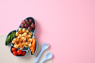 Kids meal in rocket-shaped plate with grapes, waffles, tomatoes, cucumbers, and carrots on pastel...