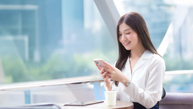 Wide 16:9 shot of young Asian businesswoman working outside office at coffee shop, using smartphone for remote communication and online business in urban city