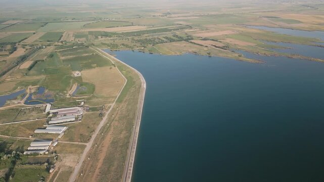 Aerial view as the camera moves above Djizak Reservoir, showing the long dam, water surface, surrounding farmlands. Roads, agricultural fields, rural buildings appear in sunlight