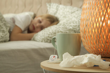 Bedside table with cup of tea, thermometer and used handchiefs and little girl sleeping in the background	

