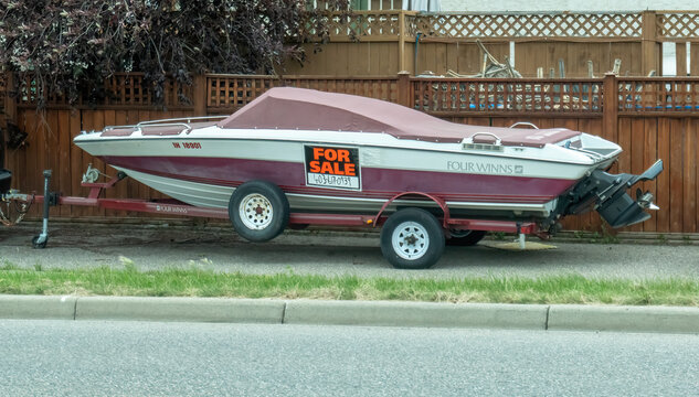 Calgary, Alberta, Canada. Jul 18, 2025. A Four Winns boat on a trailer with a "For Sale" sign is parked by a fence, covered and ready for its next owner.