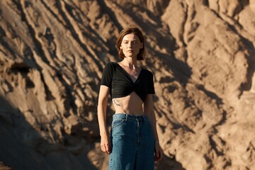 A stylish woman poses confidently in the sun against a sandy backdrop, showcasing modern fashion.