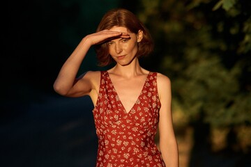 A beautiful woman shields her eyes from the sun in a red floral dress, outdoors.
