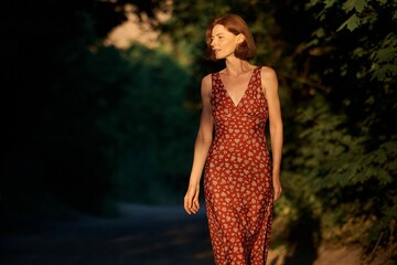 A woman strolls through a sunlit path, wearing a beautiful red floral dress.