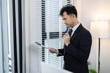 Fototapeta premium A man in a suit is standing by the window. He is talking on the phone, reading documents, and looking out the window. He checks the information in the documents.