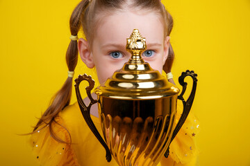 Close-up of girl with blue eyes in yellow dress hiding behind large golden trophy, playful and colorful studio shot.
