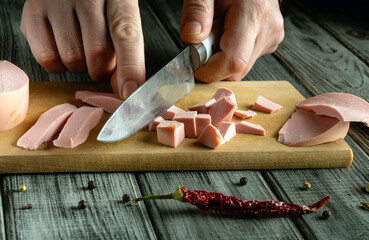 Hands expertly cut cold cuts into small cubes on a wooden board. A dried chili pepper and whole...