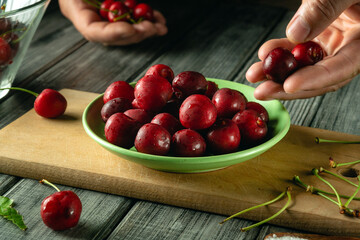 Ripe cherries are being carefully picked and placed into a green bowl on a wooden table. The warm...