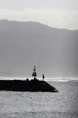 Man fishing while standing on breakwater at Haleiwa harbor fishing point, Oahu, Hawaii, USA against the light