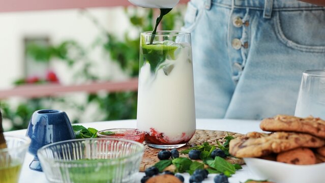 Woman pouring whisked matcha into a glass with milk, ice, and berry jam. Vibrant layered iced matcha latte preparation with fresh natural ingredients and cozy summer vibe