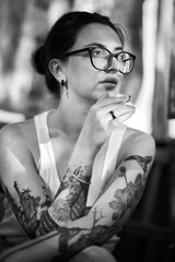 Thoughtful woman with glasses, tattoos, and a cigarette in black and white.