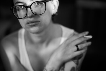 A person with glasses thoughtfully smokes a cigarette in a black and white portrait.