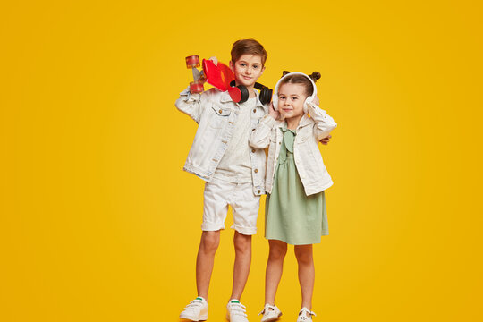 Couple of adorable stylish children with skateboard smiling and listening to music in headphones while standing against bright yellow background. Brother and sister hugging