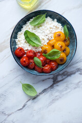 Bowl with cottage cheese, roasted tomatoes and green basil on a white stone background, vertical shot with space, above view