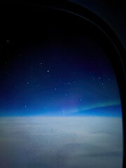 Aurora borealis northern lights seen from an airplane window at night, glowing above a blanket of clouds against a blue sky with stars