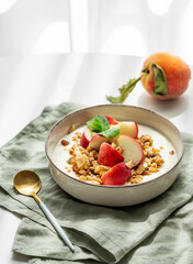 Natural greek yogurt with granola or muesli and peach in a bowl on a napkin on a white table