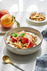 Natural greek yogurt with granola or muesli and peach in a bowl on a napkin on a white table