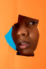 Black model peeking through torn orange paper with blue background