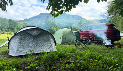 tents and red suv in green mountain valley with outdoor chairs and grill smoke rising from fire pit peaceful camping scene with hills and forest concept of summer travel adventure and outdoor living