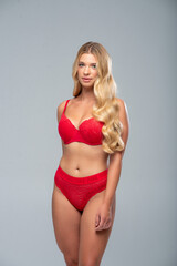 Fashion-style portrait of a slim blonde female model in red underwear, posing against a soft grey...