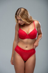 Fashion-style portrait of a slim blonde female model in red underwear, posing against a soft grey...