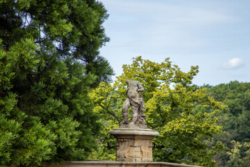 Fototapeta premium Stone statue on a pedestal surrounded by lush greenery and trees under a bright sky.