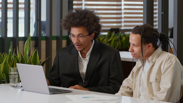 Two multiracial business men African American businessmen partners diverse colleagues males discuss online internet data analyzing marketing project result using laptop talk at office desk cooperation