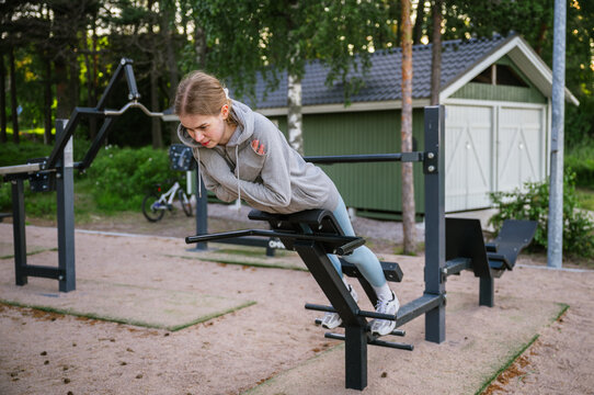Young woman doing back extension exercise on outdoor fitness bench in summer park