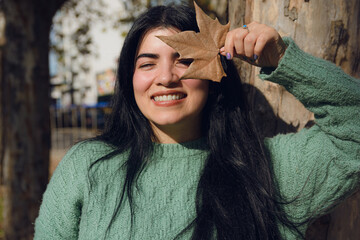 Smiling Venezuelan woman standing under the sun in the park, covering her face with a dry leaf