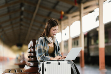 Fototapeta premium A young woman with glasses and headphones sits on a bench at a train station, holding a smartphone and looking around, surrounded by luggage and coffee.