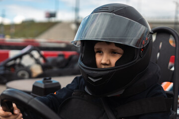 A young boy in a black protective helmet and vest drives a go-kart on a track, experiencing the...