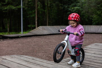 A child wearing a safety helmet and knee pads carefully rides a balance bike up a wooden ramp in a...