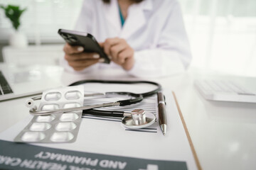 Fototapeta premium A doctor in a white coat uses a smartphone at a desk with a stethoscope, pills, medical documents, and a pen, focusing on healthcare communication and patient care.