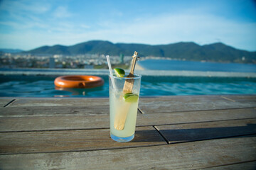 Glass of refreshing lemonade with lime and bamboo stirrer on a wooden deck by an infinity pool,...