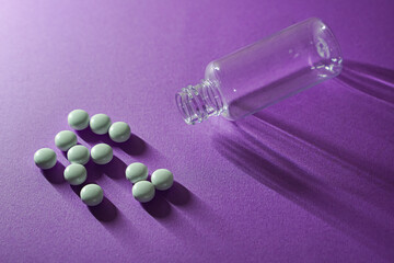 Light-gray pills scattered beside an empty plastic bottle on a purple surface.