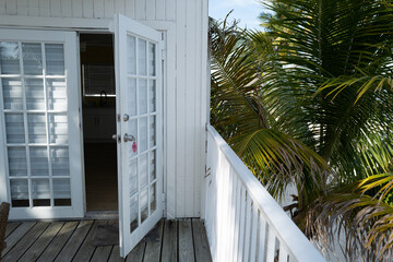 Fototapeta premium White French doors with room number 13 open onto a tropical balcony with wooden decking and lush palm leaves. Bright, airy, and inviting coastal ambiance.