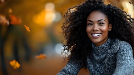 Young African American woman with curly hair wearing gray knit sweater smiling outdoors in autumn park with golden bokeh and falling leaves in background.