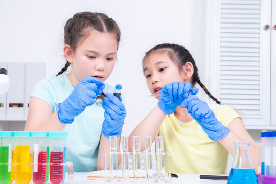 Two students wear gloves and drop colored liquid into a test tube, focus, collaborate to observe reactions in laboratory. Academic schoolgirls explore chemistry using glassware, curiosity children - Powered by Adobe
