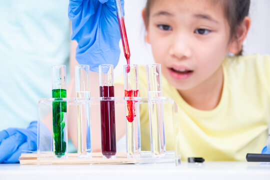Two students wear gloves and drop colored liquid into a test tube, focus, collaborate to observe reactions in laboratory. Academic schoolgirls explore chemistry using glassware, curiosity children