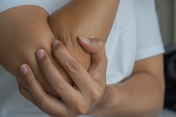 An Asian man holds his elbow, suggesting joint or tendon discomfort. His white shirt and soft-lit...