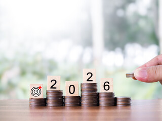 Hand putting coin into stack of coins and wooden blocks with the number 2026. The concept of saving money, Financial, Investment and Business growing in year 2026.